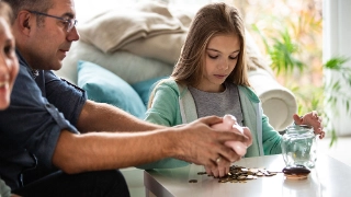 Eine Familie sitzt am Esstisch. Der Vater zaehlt gemeinsam mit seiner Tochter das Geld im Sparschwein.